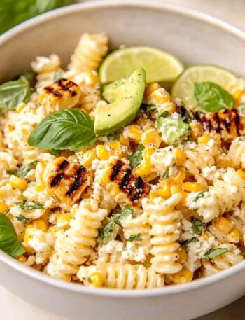 Bowl of creamy street corn pasta salad with grilled corn, rotini pasta, avocado slices, lime wedges, fresh basil, and cotija cheese