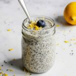 A jar of chia pudding topped with a blueberry and lemon zest, surrounded by fresh lemons and blueberries.