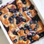 Freshly baked blueberry bread pudding topped with golden brown crust, melted cream, and juicy blueberries in a white baking dish