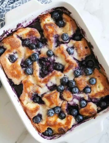Freshly baked blueberry bread pudding topped with golden brown crust, melted cream, and juicy blueberries in a white baking dish
