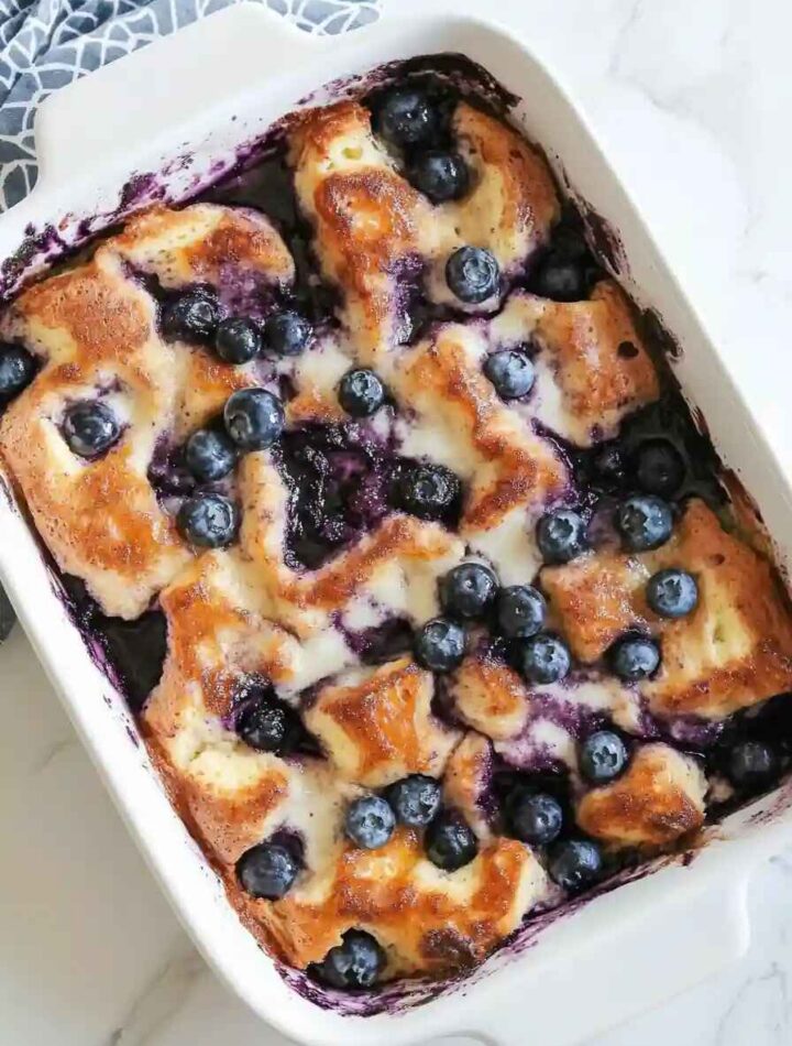 Freshly baked blueberry bread pudding topped with golden brown crust, melted cream, and juicy blueberries in a white baking dish