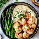 Grilled shrimp and asparagus served over creamy garlic mashed potatoes, garnished with chopped herbs in a black bowl.